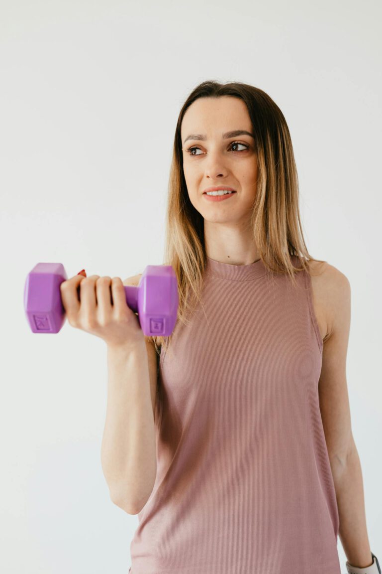 Cheerful young slim female athlete in comfy top holding dumbbell and looking away during fitness training